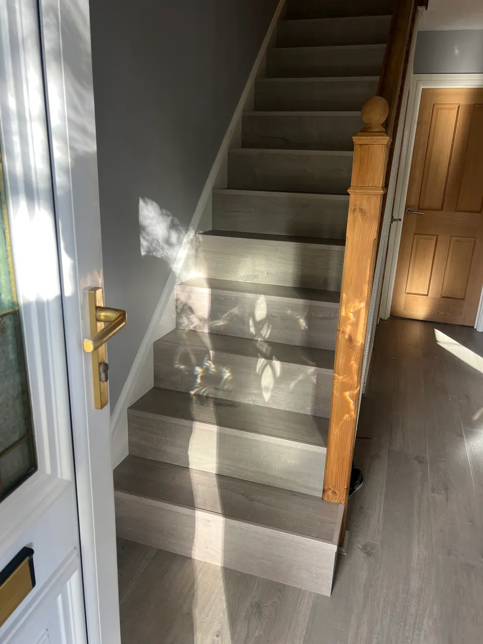 Grey laminate staircase with a pine newel post, sunlight from the front door falling across the steps